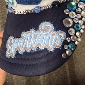 May include: A navy blue baseball cap featuring the word Spartans in light blue script with a silver glitter border. The cap is embellished with a row of light blue and silver beads, and a cluster of clear and blue rhinestones on the side panel.