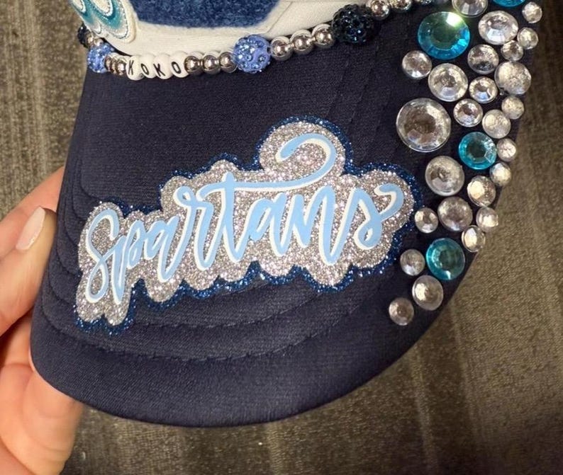 May include: A navy blue baseball cap featuring the word Spartans in light blue script with a silver glitter border. The cap is embellished with a row of light blue and silver beads, and a cluster of clear and blue rhinestones on the side panel.