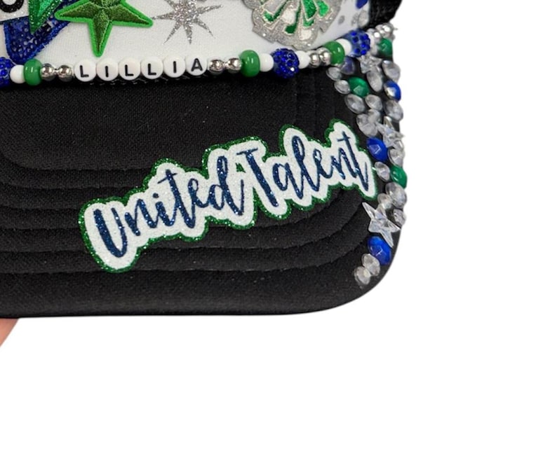 May include: A black trucker hat featuring a white front panel adorned with green star patches, silver glitter stars, and a beaded trim spelling LILLIA. The black brim displays a green and blue glitter patch with the text United Talent.