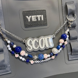 May include: A gray YETI bag with a beaded strap featuring the name "SCOTT" in white glitter. The strap includes blue beads, baseball-themed beads, and the text "GAMEDAY".