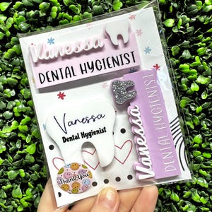 May include: A clear plastic package containing dental-themed accessories. Includes a white tooth-shaped pin, a name tag with the name "Vanessa" and the title "Dental Hygienist", and a small button with smiley faces and the words "Thank You".