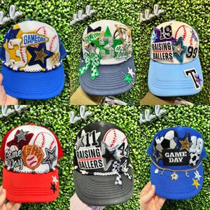 May include: Six baseball caps with assorted designs. The caps display text such as "Game Day" and "Raising Ballers", alongside baseballs, stars, and other decorations. The caps come in various colour schemes, including blue, red, and black.