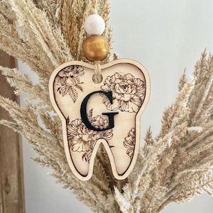 以下が含まれることがあります： 花柄と黒い「G」の文字が書かれた木製の歯のオーナメント。白いビーズと金色のビーズがついた紐で吊り下げられています。