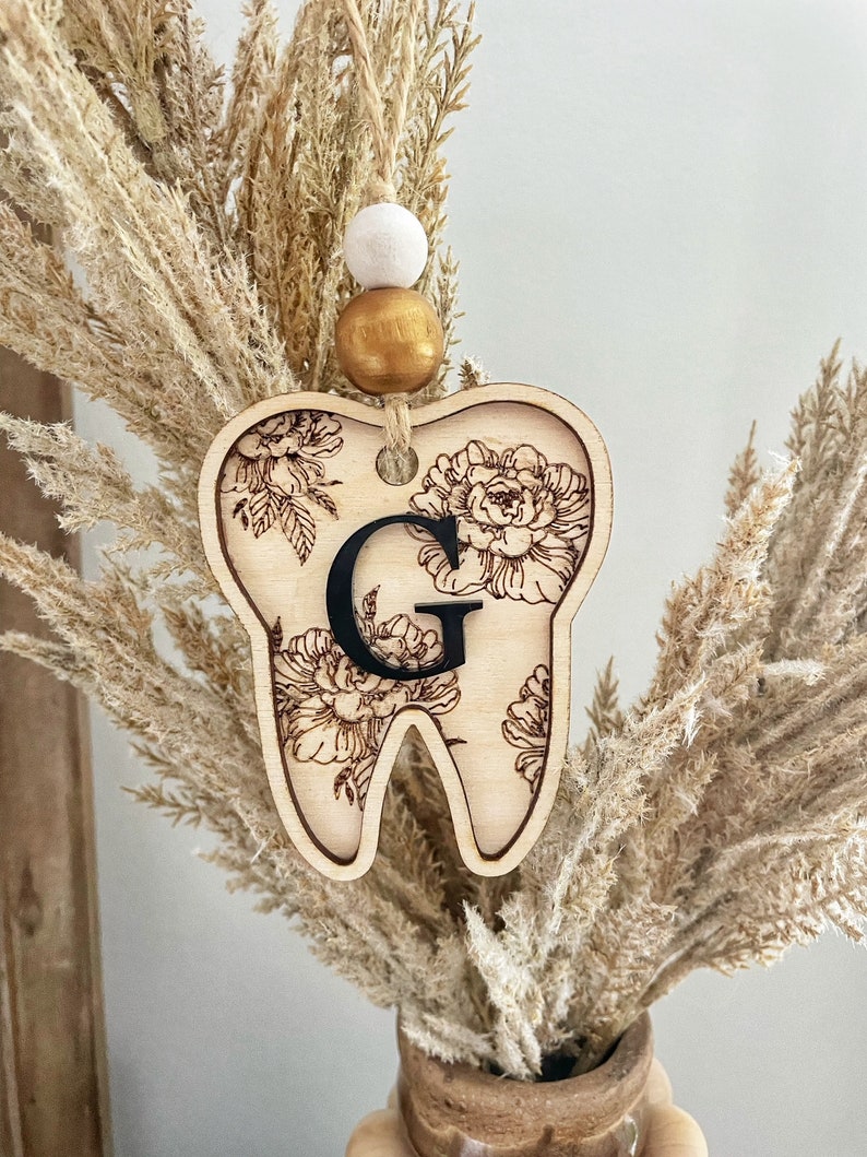 以下が含まれることがあります： 花柄と黒い「G」の文字が書かれた木製の歯のオーナメント。白いビーズと金色のビーズがついた紐で吊り下げられています。