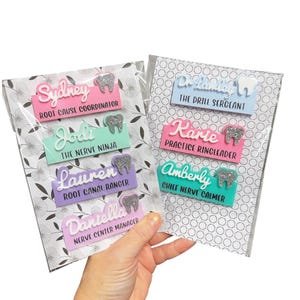 May include: A hand holds two packs of name badges. Each badge features a name, job title, and a tooth-shaped design. The badges are in various colors, including pink, blue, green, and purple, with silver glitter accents.