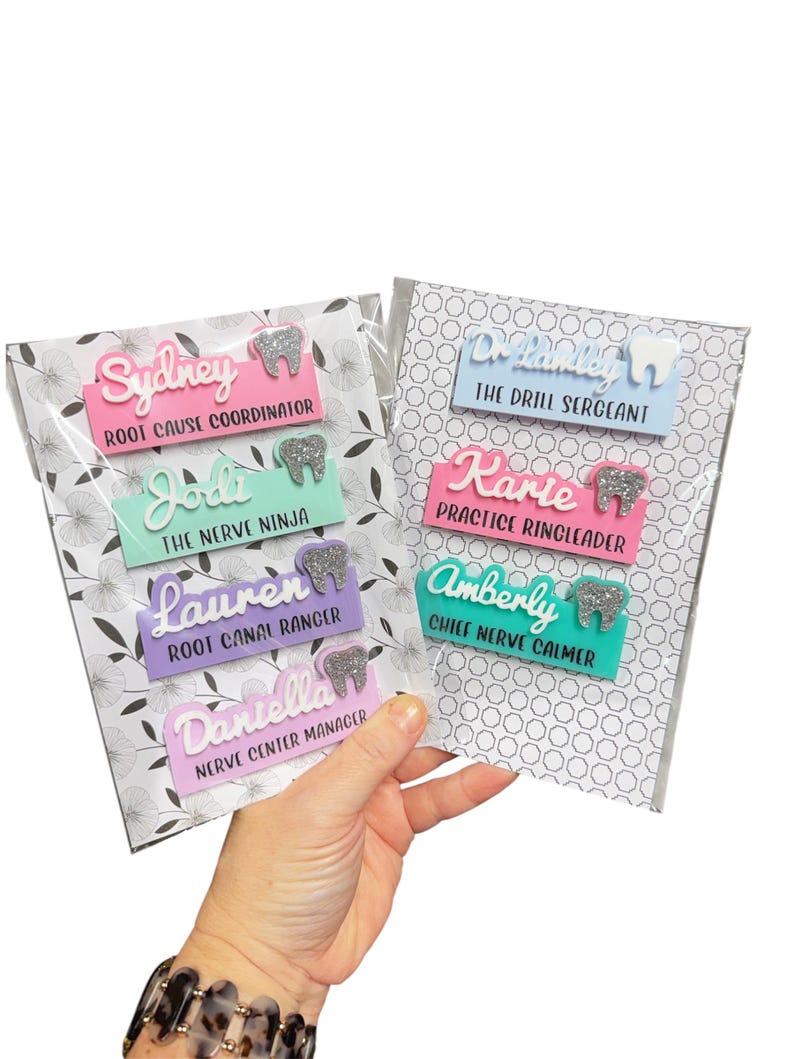 May include: A hand holds two packs of name badges. Each badge features a name, job title, and a tooth-shaped design. The badges are in various colors, including pink, blue, green, and purple, with silver glitter accents.