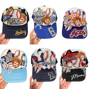 May include: Six baseball caps with baseball-themed designs. Each cap features baseballs, gloves, and decorative elements. Some caps have text like "MAMA," "Arley," "Aces," and "Cardinals." The caps are in various colours, including blue, white, red, and black.