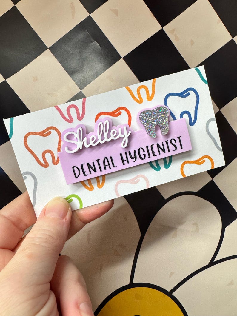 May include: A personalized name tag for a dental hygienist. The tag features the name "Shelley" in white lettering above a light purple rectangle that reads "DENTAL HYGIENIST". A glittery tooth-shaped embellishment is on the right, and the background is a pattern of colorful teeth.