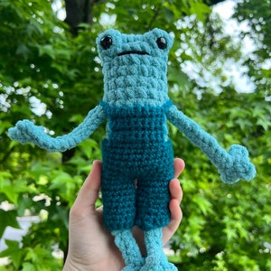 May include: A crocheted blue frog wearing blue overalls. The frog is holding its arms out to the sides.