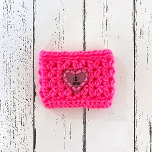 May include: A pink crocheted cup cosy with a heart-shaped leather patch that says "mine" in black lettering.