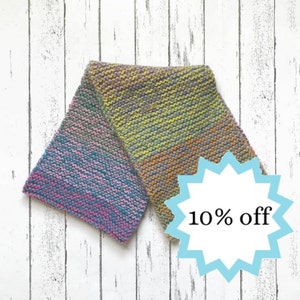 May include: A colorful knitted scarf with a gradient of pink, blue, green, and yellow. The scarf is folded in half and displayed on a white wooden background. A blue starburst with the text "10% off" is in the lower right corner of the image.