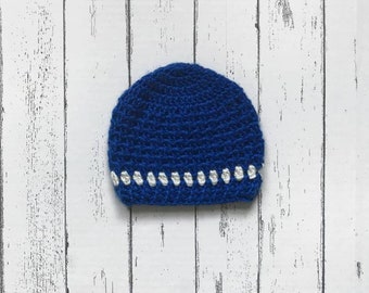 May include: A blue crocheted hat with a white crocheted trim. The hat is on a white wooden background.