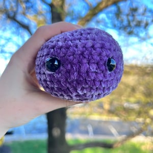 May include: A purple crocheted plush toy with black eyes. The toy is being held in a hand.