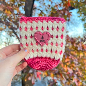 May include: A handmade crochet cup cozy in white and pink. The cozy features a diamond pattern with a pink heart-shaped patch that says "mine". The cozy is designed to fit a standard-sized cup.