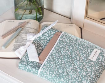 May include: A green floral-patterned quilted pouch with a white ribbon and a light brown object peeking out. Two rectangular objects, one light brown and one light blue, are next to an open book. A vase of flowers sits in the background.