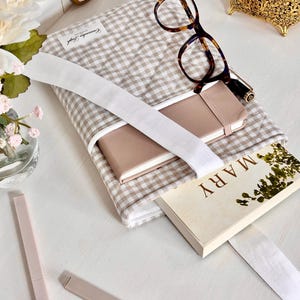 May include: A beige and white gingham book sleeve with a white strap, holding a pink notebook and a book with the title "MARY". Tortoiseshell glasses and a gold jewellery box are also on the white surface. Two pink cosmetic tubes are also visible.