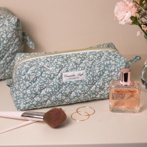 Peut inclure: Une trousse de maquillage matelassée à motifs floraux, de couleurs bleu sarcelle et blanc, avec une fermeture éclair dorée. L'étiquette indique "Cassandra Leigh Handmade". Un pinceau de maquillage, deux bagues dorées et un flacon de parfum sont également visibles.