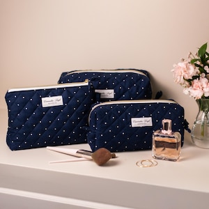 Peut inclure: Trois trousses de maquillage matelassées bleu marine à pois blancs et fermetures éclair dorées. Les sacs sont disposés sur une surface blanche avec des pinceaux de maquillage, du parfum et des fleurs en arrière-plan. Chaque sac a une étiquette blanche avec du texte noir.
