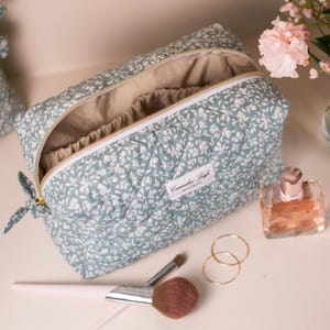May include: A floral-patterned quilted cosmetic bag in blue and white, with a beige interior. The bag is open, revealing the inside. A perfume bottle, makeup brushes, and gold hoop earrings are also visible.