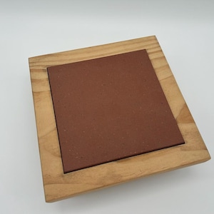 May include: A square terracotta tile resting on a wooden frame. The tile is a reddish-brown color and has a rough texture. The frame is made of light brown wood and has a simple design.
