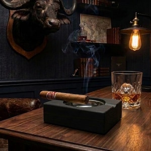 May include: A lit cigar rests in a black ashtray on a wooden desk. A glass of whiskey, a vintage lamp, and a leather chair are also visible. A buffalo head mount hangs on the dark blue wall, creating a masculine, vintage aesthetic.
