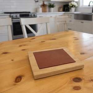 May include: A square wooden coaster with a reddish-brown center sits on a light-colored wooden table. The coaster has a light wood frame. The table is made of light-colored wood with visible knots.