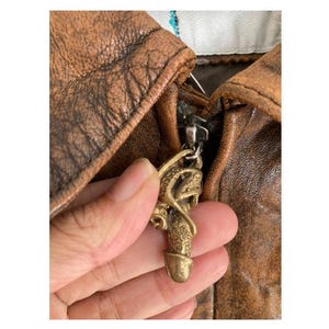 Op de afbeelding: Close-up van een bruine leren jas met een metalen rits en een decoratieve ritstrekker. De ritstrekker is een goudkleurige, fallusvormige bedel met ingewikkelde details. Het leer van de jas heeft een distressed uitstraling.