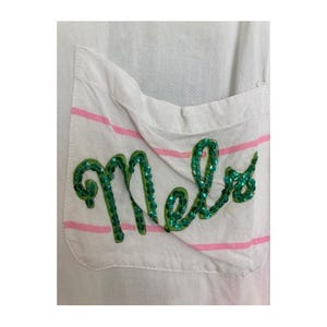 Puede incluir: Primer plano de un bolsillo de tela blanca con rayas horizontales rosas. La palabra "Mels" está bordada con lentejuelas verdes. El bolsillo está adherido a una prenda blanca.