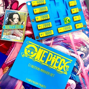 May include: A blue One Piece condition marker set with yellow numerical tokens, a green die, and a card featuring Boa Hancock. The set includes a blue storage box with the text One Piece, Condition Marker Set, and Phosphory Prints printed in yellow.