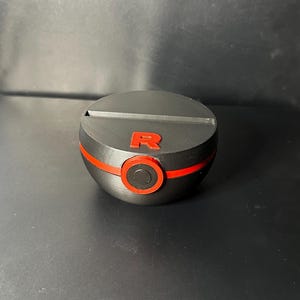 Peut inclure: Un support de Poké Ball imprimé en 3D, noir et rouge. Le support présente une bande rouge au milieu et un cercle rouge avec un petit motif. Le dessus porte un logo "R" rouge.