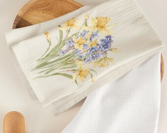 May include: A white kitchen towel with a floral design featuring yellow daffodils and blue hyacinths. The towel is folded and displayed on a wooden surface.