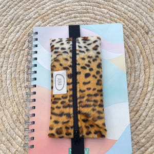 Könnte beinhalten: Ein Spiralnotizbuch mit farbenfrohem Einband wird mit einem Federmäppchen mit Leopardenmuster kombiniert. Das Federmäppchen hat einen schwarzen Reißverschluss und ein schwarzes Gummiband. Ein kleines weißes Etikett auf dem Federmäppchen trägt die Aufschrift "ALMA".