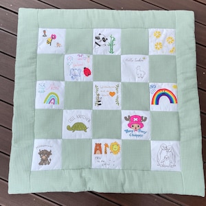 Könnte beinhalten: Eine hellgrüne Babydecke mit Waffelstruktur. Die Decke hat bestickte Quadrate mit verschiedenen Designs, darunter ein Regenbogen, eine Schildkröte, ein Panda und der Text "Hello Sunshine".