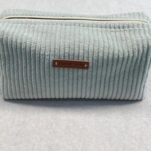 Op de afbeelding: Een rechthoekige, lichtblauwe corduroy toilettas met een witte rits en een klein bruin "Handmade" label. De tas heeft een zacht, getextureerd uiterlijk en een eenvoudig, klassiek ontwerp.