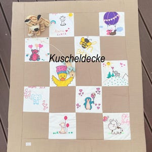 May include: A beige waffle-textured blanket featuring a patchwork design of white squares with colourful illustrations of animals, balloons, and the text "Kuscheldecke," "Aseria," and "Oh Baby." A small plush tiger and a wooden toy are attached.