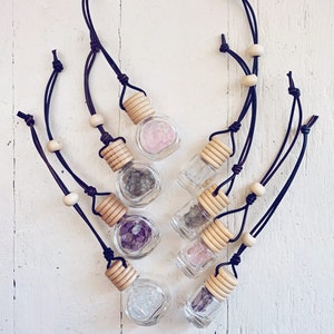 May include: Several small glass bottles filled with gemstones, such as rose quartz and amethyst. Each bottle has a wooden cap and is suspended by a dark brown leather cord with wooden beads. The bottles are displayed on a white surface.