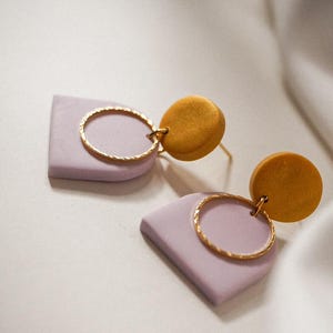 Könnte beinhalten: Ein Paar handgefertigte Ohrringe mit geometrischem Design. Jeder Ohrring hat eine lavendelfarbene, abgerundete quadratische Form, einen goldfarbenen Ring und einen senfgelben Kreis. Die Ohrringe werden auf einer weißen Oberfläche präsentiert.
