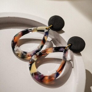 May include: A pair of teardrop-shaped earrings with a marbled pattern in shades of orange, purple, yellow, and white. The earrings have a black circular stud and gold-colored hooks. The earrings are displayed on a white surface.