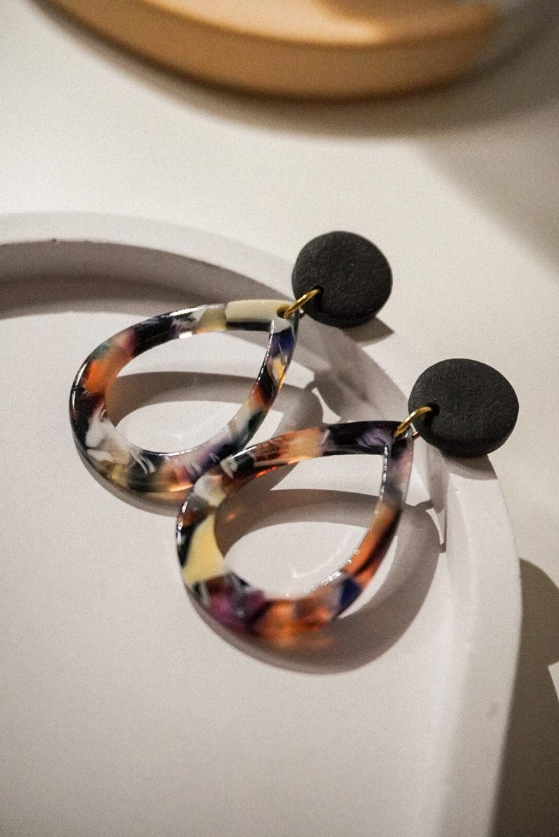 May include: A pair of teardrop-shaped earrings with a marbled pattern in shades of orange, purple, yellow, and white. The earrings have a black circular stud and gold-colored hooks. The earrings are displayed on a white surface.