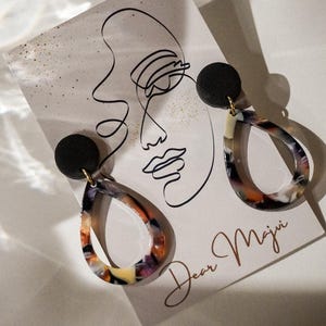 May include: A pair of teardrop earrings with a black stud and a colorful, marbled design in shades of orange, purple, and cream. The earrings are displayed on a card with a line drawing of a face and the text "Dear Maji".