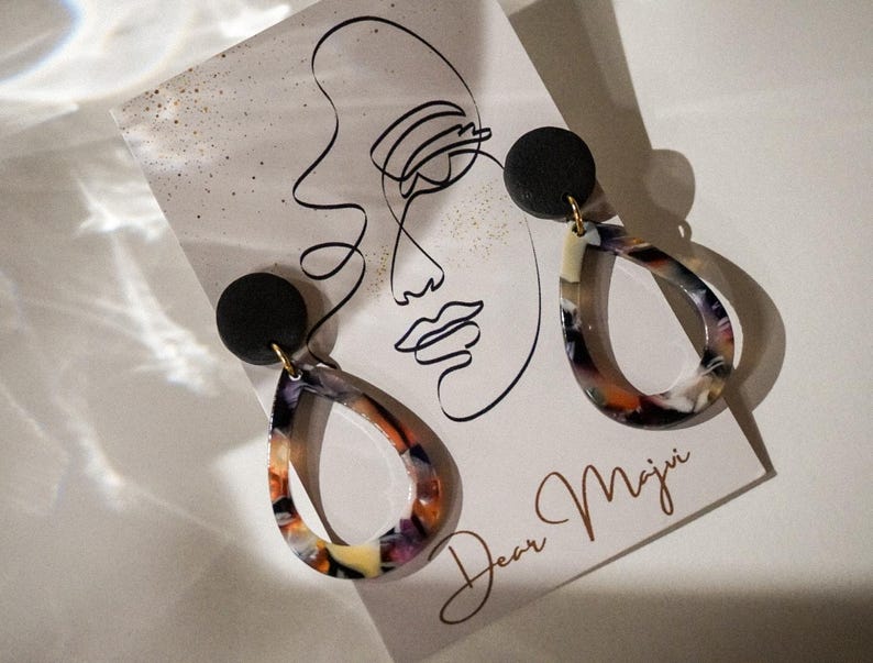 May include: A pair of teardrop earrings with a black stud and a colorful, marbled design in shades of orange, purple, and cream. The earrings are displayed on a card with a line drawing of a face and the text "Dear Maji".
