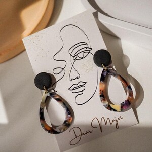 May include: A pair of teardrop earrings with a black stud top and a colorful, marbled design. The earrings are displayed on a card with a line drawing of a face and the text "Dear Maju".