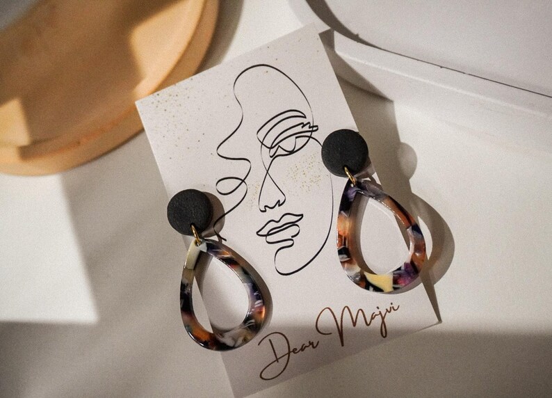 May include: A pair of teardrop earrings with a black stud top and a colorful, marbled design. The earrings are displayed on a card with a line drawing of a face and the text "Dear Maju".