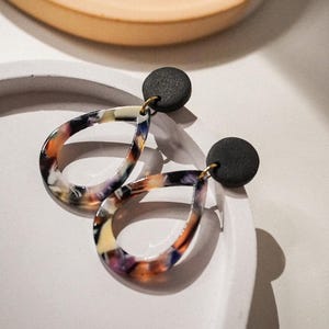 May include: A pair of teardrop-shaped earrings with a marbled pattern in shades of orange, purple, and white. The earrings have a black circular stud and gold-colored hooks. The earrings are displayed on a white surface.