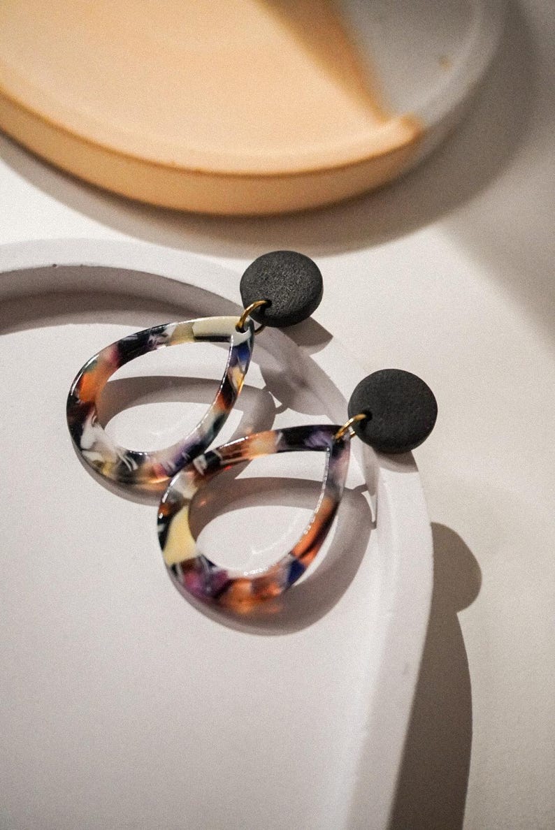 May include: A pair of teardrop-shaped earrings with a marbled pattern in shades of orange, purple, and white. The earrings have a black circular stud and gold-colored hooks. The earrings are displayed on a white surface.