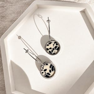 以下が含まれることがあります： クリーム色のベースに不規則な黒い斑点模様が入った円形のペンダントが付いた、シルバーのフープピアス一対。このピアスは、細いシルバーワイヤーのフープデザインで、平らな円形の模様入りディスクパーツに接続されています。