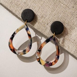 May include: A pair of teardrop earrings with a black circular stud and a colorful, patterned teardrop pendant. The pendant features a mix of orange, yellow, purple, and black hues. The earrings are suspended by a gold-colored hook.