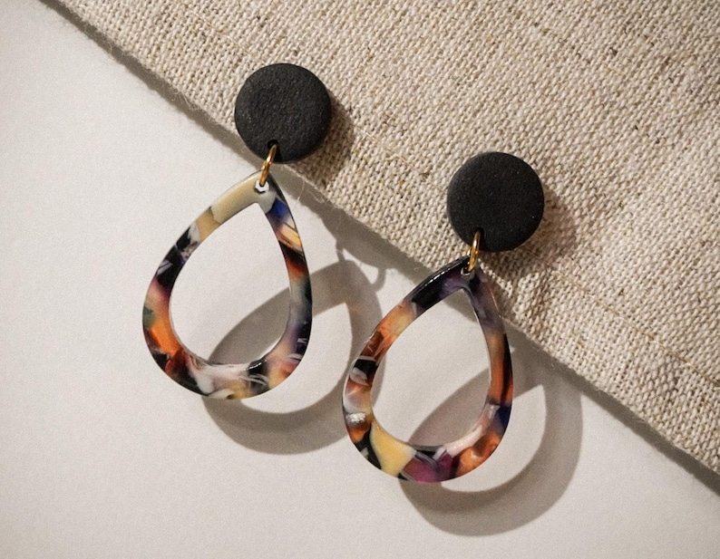 May include: A pair of teardrop earrings with a black circular stud and a colorful, patterned teardrop pendant. The pendant features a mix of orange, yellow, purple, and black hues. The earrings are suspended by a gold-colored hook.