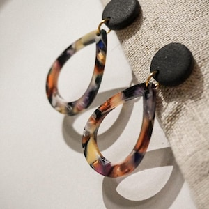 May include: A pair of teardrop earrings with a black circular stud and a colorful, marbled design. The earrings have a gold-colored hook and are set against a neutral background.