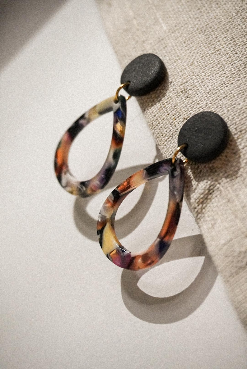 May include: A pair of teardrop earrings with a black circular stud and a colorful, marbled design. The earrings have a gold-colored hook and are set against a neutral background.